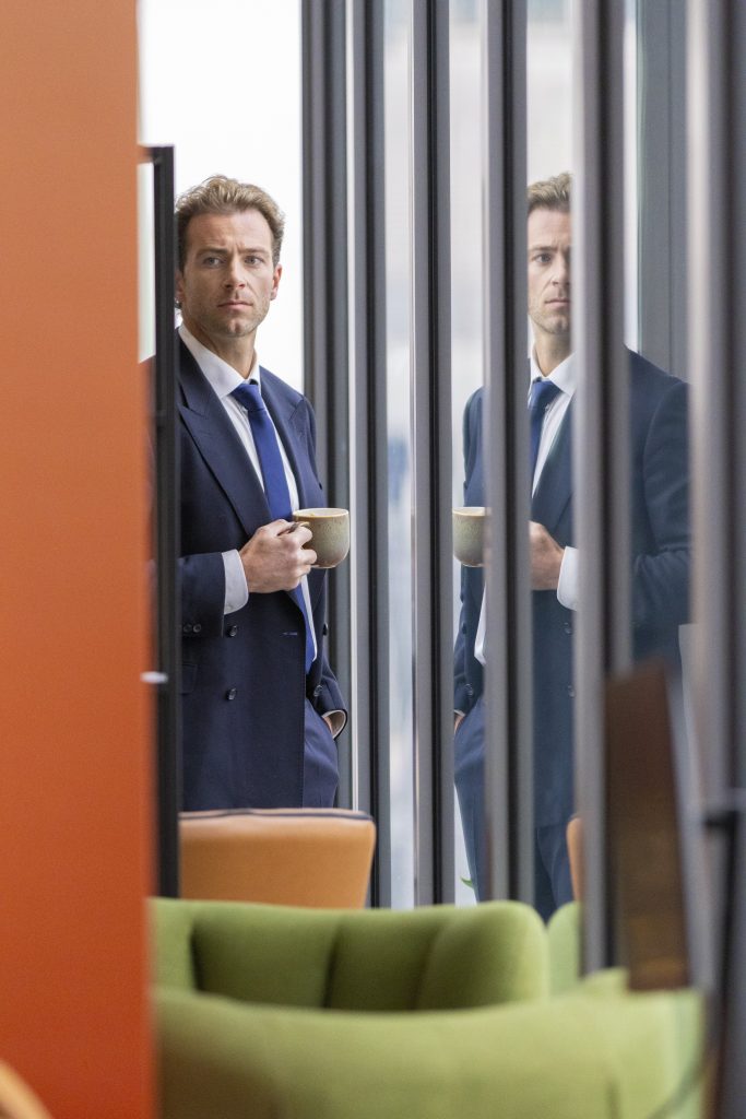 a man in a navy suit stands opposite floor-to-ceiling window in an office building. An orange wall is to his left and green sofa chairs sit in front of the window.