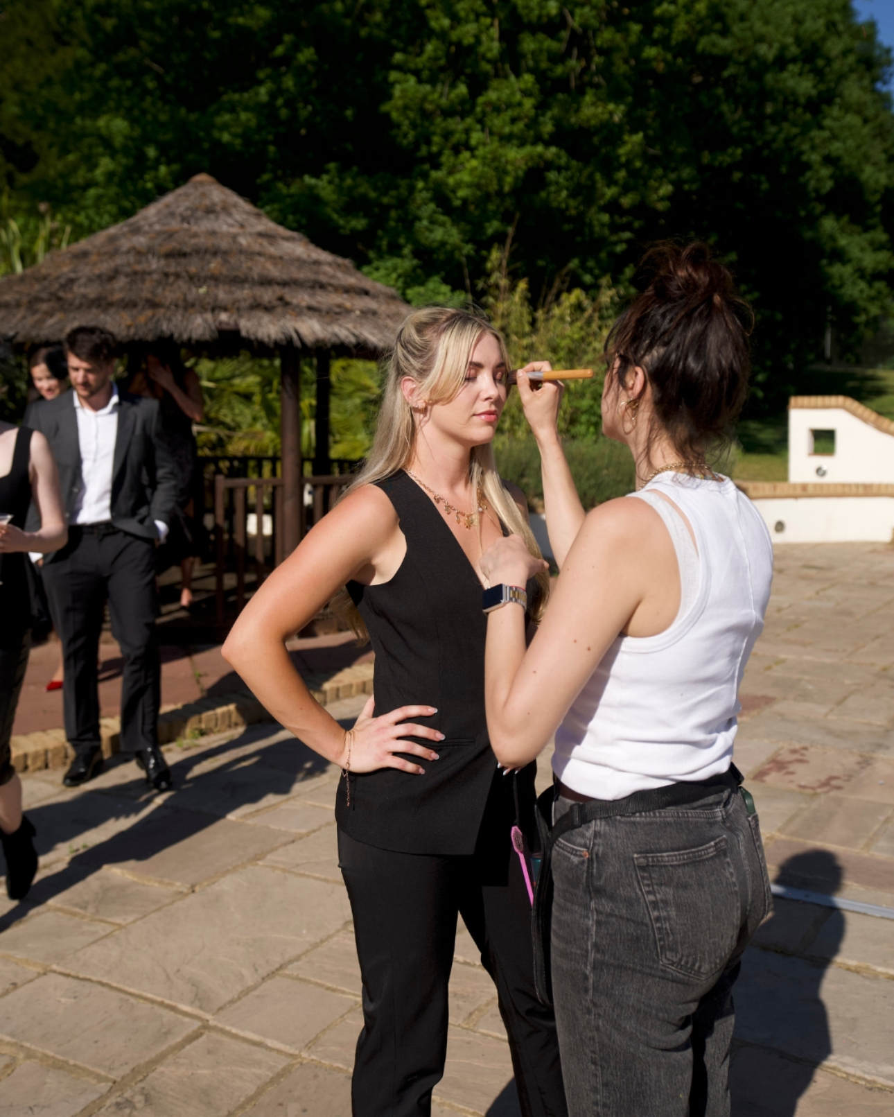 makeup-artist-applying-makeup A woman with brown hair tied up wearing a white vest and grey trousers has her back to the camera. She applies makeup to a blonde woman in a black jumpsuit. They are stood in a large garden with a stone patio and tiki hut in the background.