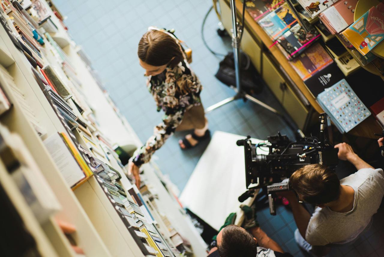 Birds eye shot of a film shoot in a library. A woman stands picking a book off of a shelf. A C-stand is behind her and a camera point at her face.