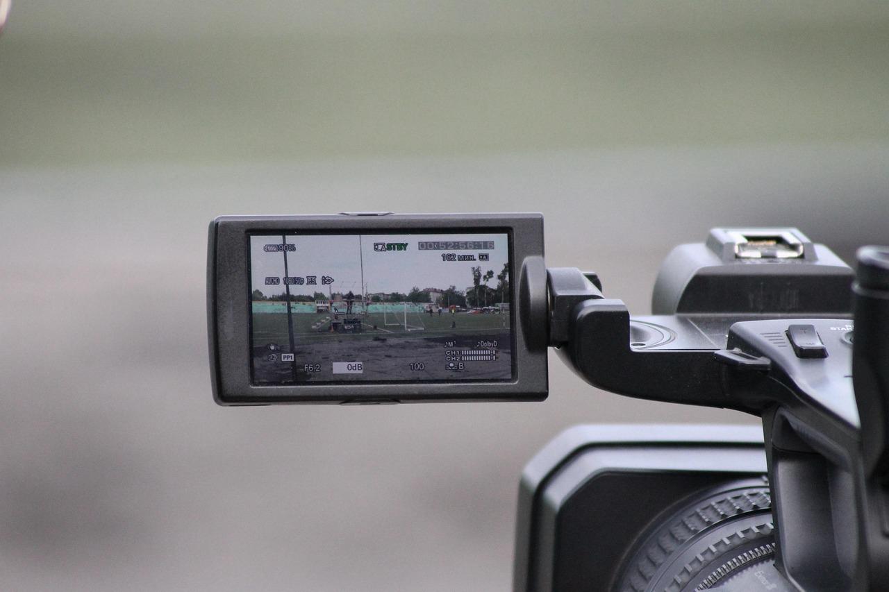 behind-a-video-camera POV from behind a large video camera. We can see the body of the camera on the right and the screen showing the display on the left. The screen shows a green landscape.