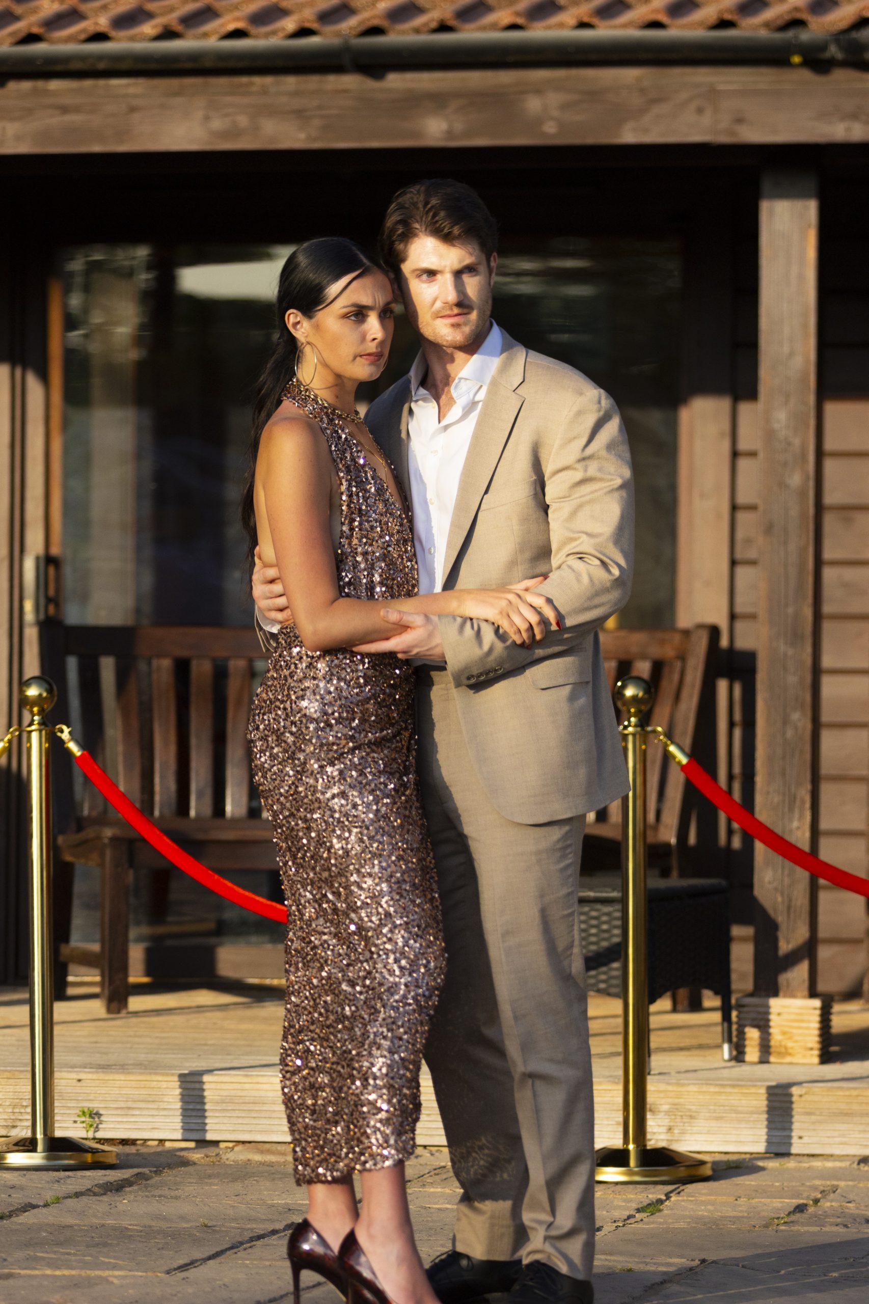 Two glamorous actors on a vertical drama set. On the left there is a tanned woman in a sparkly gold dress, on the right a suave man in a cream suit.