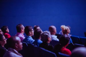 An auditorium with an audience. They are lit by a blue almost LED light. We can see the back of their heads.