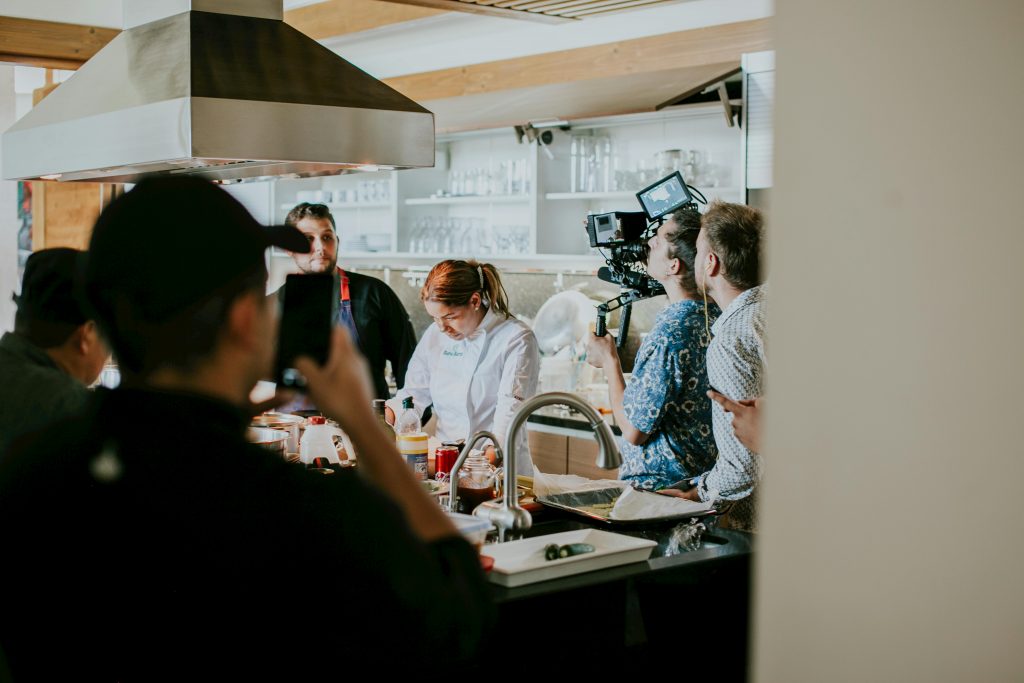 A film crew stood around a bright white kitchen table. A chef stands in the middle with a pin stripe apron on. Cameras pointed at her.