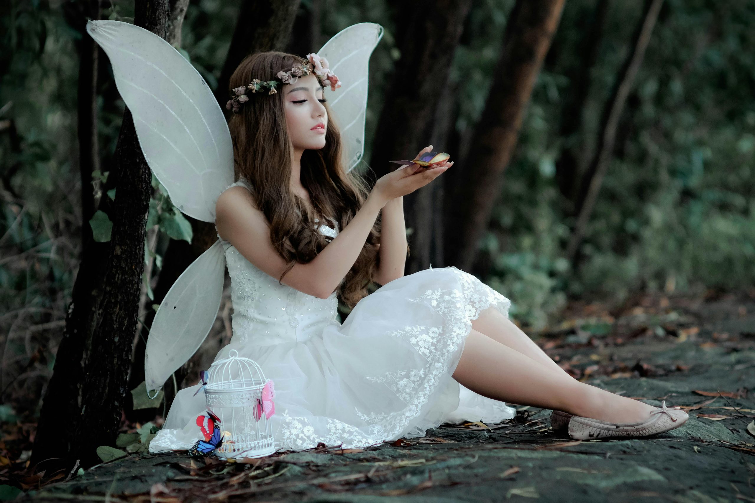Young woman wearing a white dress with white wings sits down against a tree in the forrest. She wears a flower crown of pink flowers and holds a butterfly in her hands.