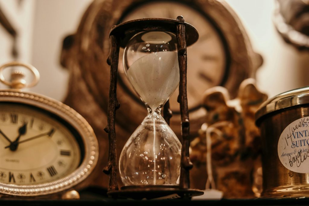 A glass sand timer with wooden top bottom and pillars is in the centre of the frame surrounded by brass vintage clocks. The picture represents the passing of time.