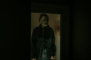 Mike Myers from the 'Halloween' film series. He stands inside a dimly lit doorway.