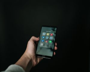 An iPhone held in a white hand wearing a grey jumper. The apps are inside a 'social media' folder.