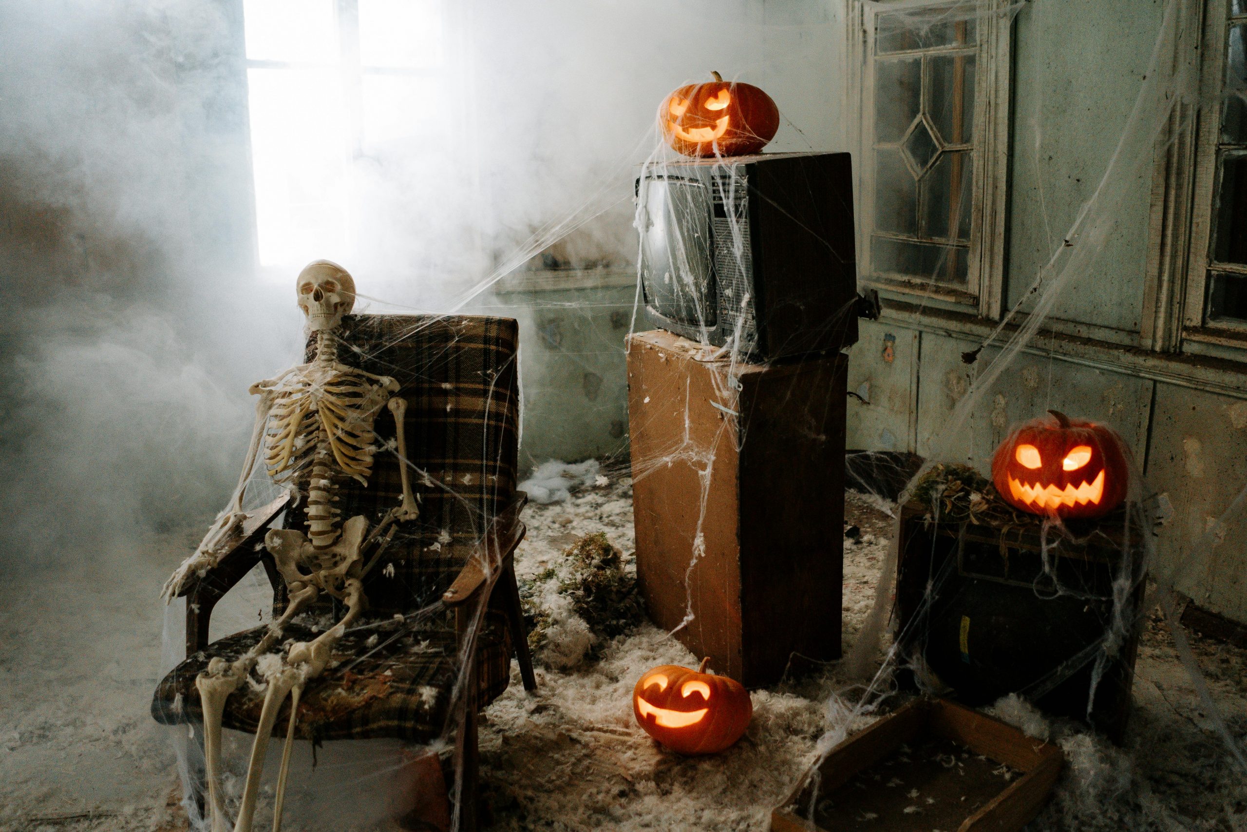 Skeleton sits in a creepy haunted house. Surrounded by cobwebs and lit pumpkins. He sits in front of a vintage television.