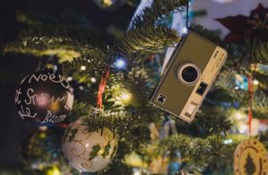 A close up of a Christmas tree with a camera ornament, the tree is full of other brightly covered ornaments.